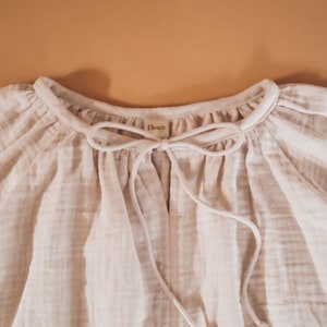 Pode incluir: Uma blusa branca de algod&atilde;o com decote franzido e um la&ccedil;o na frente. A blusa tem uma pequena etiqueta que diz "Flora" na frente.
