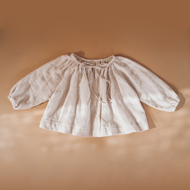 Pode incluir: Uma blusa branca de algod&atilde;o em gaze estilo camponesa com mangas compridas e um la&ccedil;o no decote.