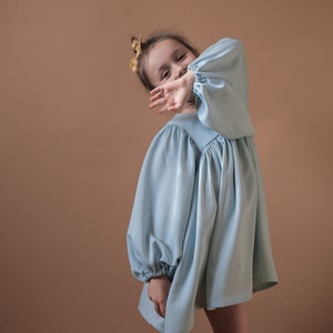 Könnte beinhalten: Ein junges Mädchen trägt ein hellblaues Kleid mit Puffärmeln und einer gelben und weißen karierten Schleife im Haar. Sie lächelt und bedeckt ihr Gesicht mit der Hand.
