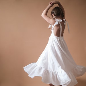 Könnte beinhalten: Ein junges Mädchen trägt ein weißes Kleid aus Spitze mit tiefem Rücken und Schleifenbändern. Das Kleid hat einen Rüschen-Saum und weht im Wind.