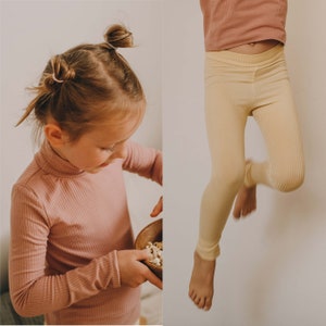 Könnte beinhalten: Zwei Fotos von Kindern, die gerippte Strickoberteile tragen. Das erste Foto zeigt ein Kind, das ein rosa geripptes Rollkragenoberteil trägt. Das zweite Foto zeigt ein Kind, das ein gelbes geripptes Oberteil trägt und in die Luft springt.