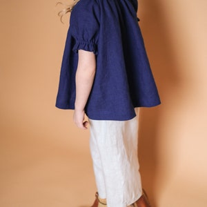 Pode incluir: Uma pessoa jovem vestindo uma blusa de linho azul marinho com mangas bufantes e cal&ccedil;as de linho brancas. Ela est&aacute; usando botas de couro marrom com um detalhe dourado.