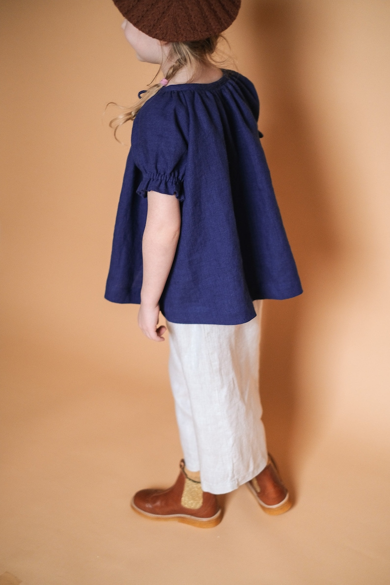 Pode incluir: Uma pessoa jovem vestindo uma blusa de linho azul marinho com mangas bufantes e cal&ccedil;as de linho brancas. Ela est&aacute; usando botas de couro marrom com um detalhe dourado.