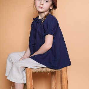 Pode incluir: Uma menina jovem vestindo uma blusa azul com um beret marrom est&aacute; sentada em um banquinho de madeira. Ela est&aacute; vestindo cal&ccedil;as brancas e botas marrons.