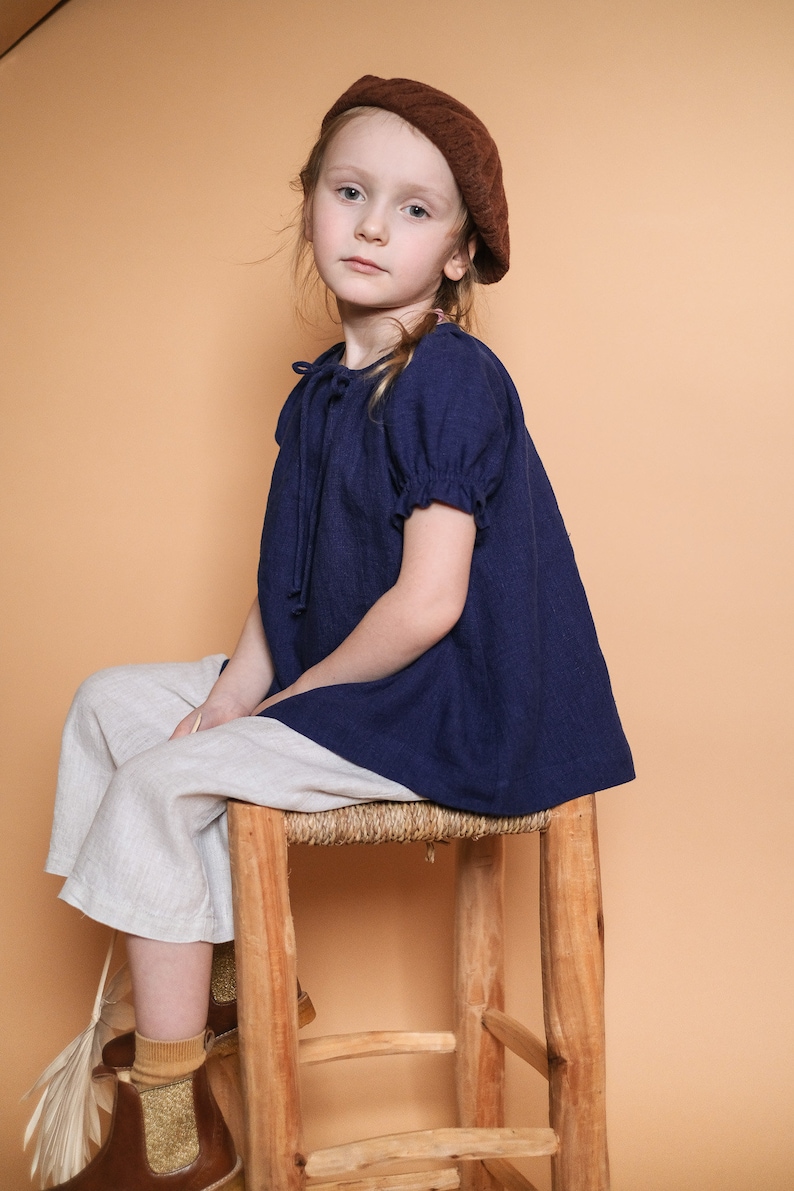 Pode incluir: Uma menina jovem vestindo uma blusa azul com um beret marrom est&aacute; sentada em um banquinho de madeira. Ela est&aacute; vestindo cal&ccedil;as brancas e botas marrons.