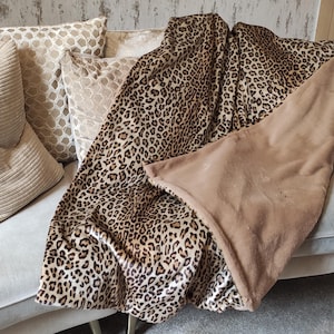 May include: A leopard print blanket on a light-coloured sofa with several decorative cushions. The blanket has a brown and black spotted pattern on one side and a plain tan colour on the other. The cushions are in neutral tones.