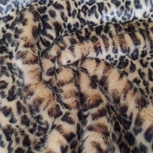Puede incluir: Primer plano de una tela de piel sint&eacute;tica con estampado de leopardo. La tela presenta una base beige con manchas marrones oscuras y negras, creando un dise&ntilde;o texturizado y animal. El material parece suave y afelpado.
