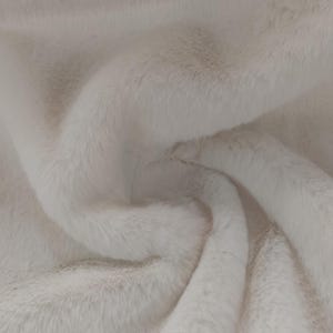 May include: Close-up of a soft, off-white faux fur fabric. The plush material has a dense pile, with long, flowing fibers creating a textured, luxurious appearance. The fabric is gently folded, showing the depth and dimension of the material.