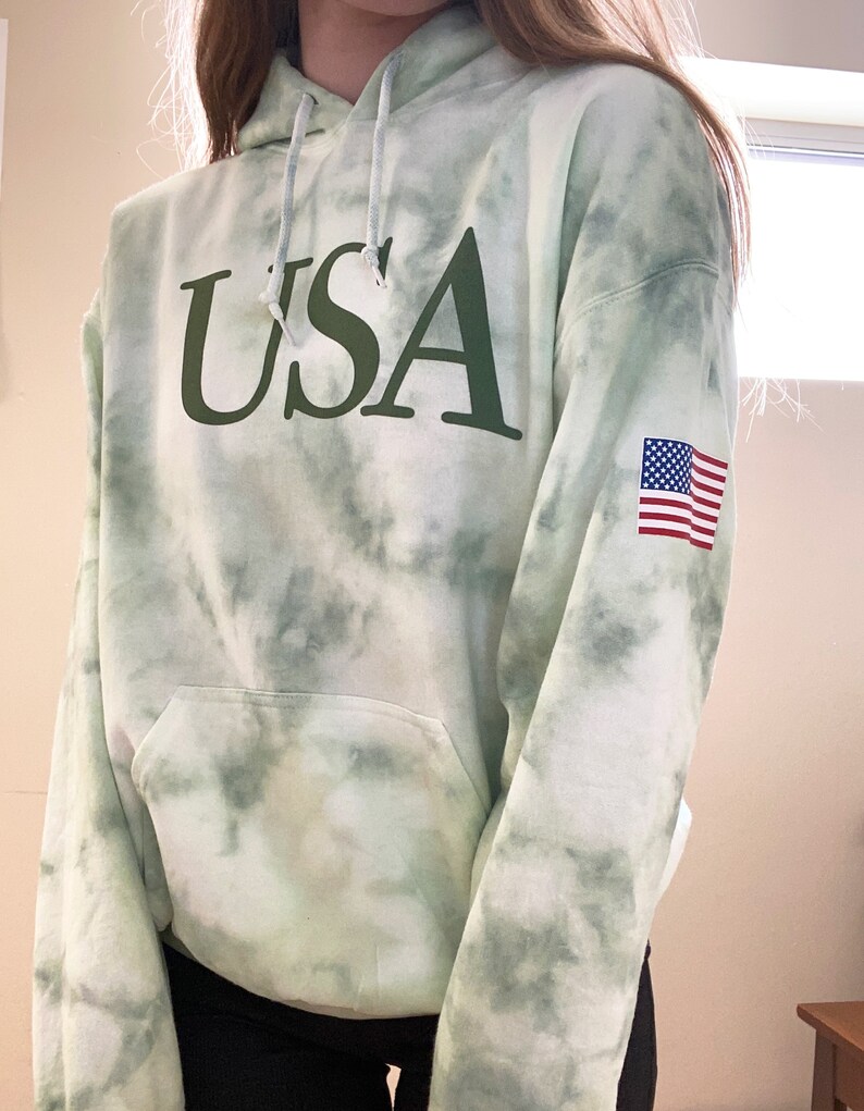 Pu&ograve; includere: Felpa con cappuccio verde tie-dye con la scritta "USA" stampata in verde sul davanti e una piccola toppa della bandiera americana sulla manica.