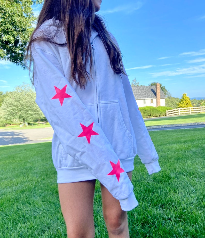 May include: White zippered hoodie with pink stars on the sleeves. The hoodie is made of a soft, comfortable material.