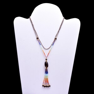 Pode incluir: Um colar de várias voltas com um pingente de pedra marrom e um tassel de contas coloridas. O colar é feito de pequenas contas facetadas em tons de azul, verde, laranja e vermelho.