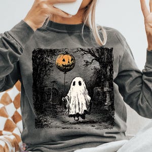 May include: Dark grey long-sleeved shirt with a Halloween graphic. The graphic shows a ghost holding a pumpkin balloon in a graveyard. The shirt is worn with grey sweatpants and a white beanie hat.