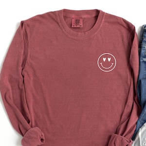 May include: A maroon long-sleeved t-shirt with a white smiley face with heart eyes graphic on the left chest.