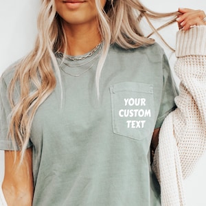 Op de afbeelding: Een saliegroen T-shirt met een zakje aan de linkerkant. Het zakje heeft witte tekst met de tekst "YOUR CUSTOM TEXT".