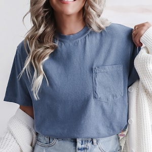 Op de afbeelding: Een blauw T-shirt met korte mouwen en een borstzak. Het shirt is effen van kleur en heeft een relaxte pasvorm. De woorden "CUSTOM DESIGN" zijn in het zwart op een witte achtergrond gedrukt.