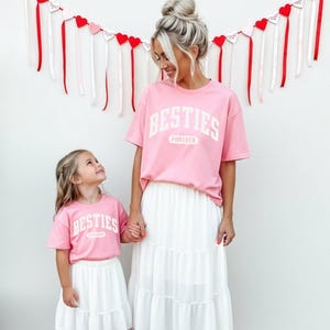 May include: Matching pink t-shirts with the word "BESTIES" in white block letters, paired with white tiered skirts. The shirts are worn by a woman and a young girl, holding hands. Red and white heart decorations hang in the background.