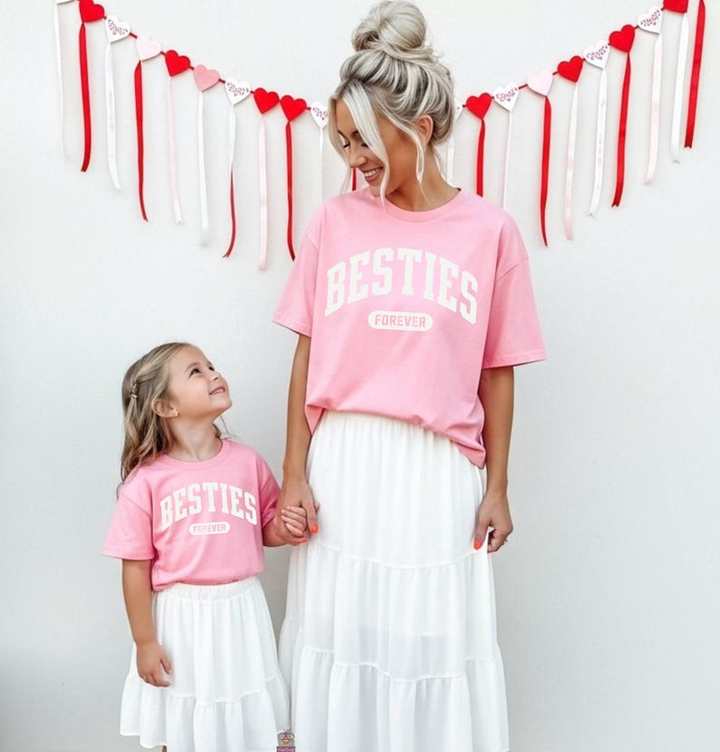 May include: Matching pink t-shirts with the word "BESTIES" in white block letters, paired with white tiered skirts. The shirts are worn by a woman and a young girl, holding hands. Red and white heart decorations hang in the background.
