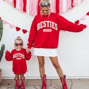 May include: Matching red sweatshirts with the text "BESTIES FOREVER" in white. The sweatshirts are worn by a woman and a child, paired with red cowboy boots. The background features Valentine's Day decorations.