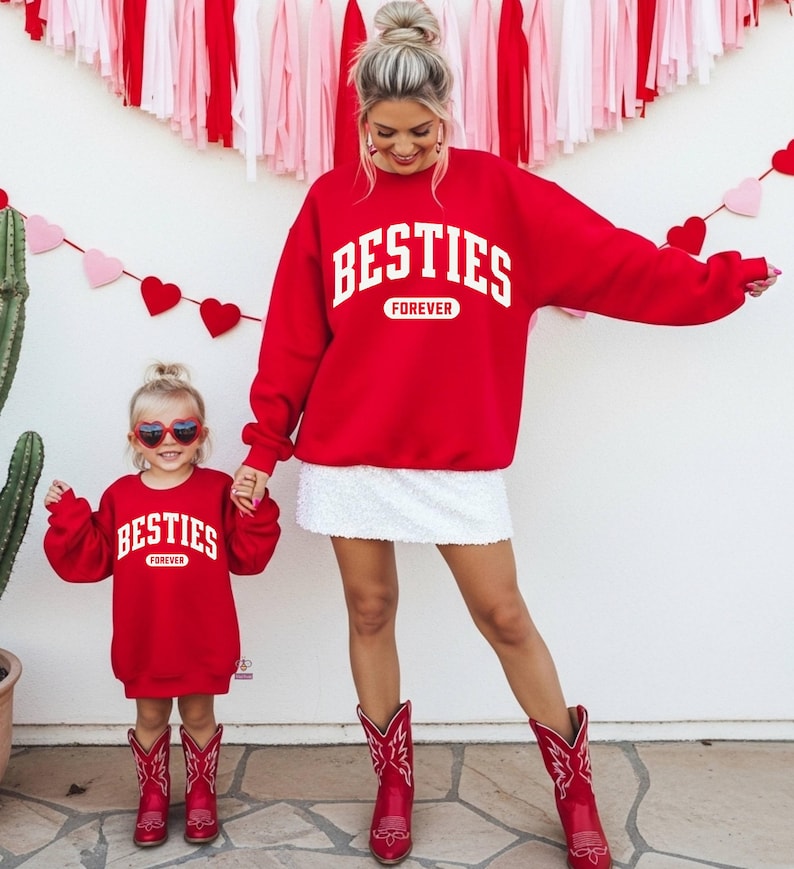 May include: Matching red sweatshirts with the text "BESTIES FOREVER" in white. The sweatshirts are worn by a woman and a child, paired with red cowboy boots. The background features Valentine's Day decorations.