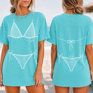 May include: A turquoise short-sleeve t-shirt featuring a white line-art graphic of a bikini. The front displays the bikini top and bottom with the text 'East Hampton 2024'. The back shows the bikini bottom and the tie-back detail of the top.