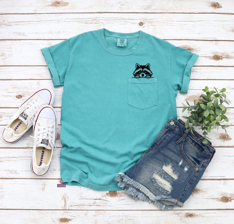 May include: A turquoise blue t-shirt with a front pocket featuring a black and white raccoon peeking out. The t-shirt has short sleeves and a crew neck.