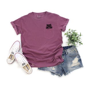 May include: A mauve t-shirt with a front pocket featuring an embroidered raccoon peeking out. The t-shirt has rolled up sleeves and is paired with denim shorts.