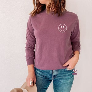 May include: A long-sleeved, mauve colored t-shirt with a white smiley face graphic with heart eyes.