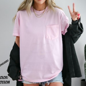 Może przedstawiać: Jasnor&oacute;żowy t-shirt z kr&oacute;tkim rękawem i kieszenią na piersi po lewej stronie. Koszulka jest noszona z czarną koszulą z długim rękawem i warstwowymi złotymi naszyjnikami. Tekst na obrazku brzmi: PERSONALIZABLE COLOR: BLOSSOM.