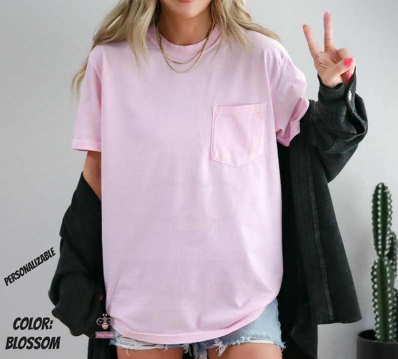 Może przedstawiać: Jasnor&oacute;żowy t-shirt z kr&oacute;tkim rękawem i kieszenią na piersi po lewej stronie. Koszulka jest noszona z czarną koszulą z długim rękawem i warstwowymi złotymi naszyjnikami. Tekst na obrazku brzmi: PERSONALIZABLE COLOR: BLOSSOM.