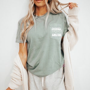 May include: A sage green t-shirt with a white pocket featuring the text "FUTURE MRS. SMITH" in a retro font.