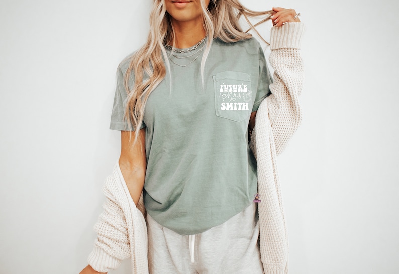May include: A sage green t-shirt with a white pocket featuring the text "FUTURE MRS. SMITH" in a retro font.