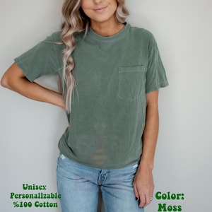 Może przedstawiać: T-shirt z kr&oacute;tkim rękawem w kolorze mchu z okrągłym dekoltem i kieszenią na piersi po lewej stronie. Koszulka jest unisex i wykonana w 100% z bawełny. Tekst na obrazku brzmi: Unisex, Personalizable, %100 Cotton, Color: Moss.