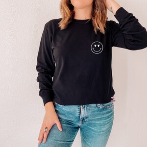 May include: A black long-sleeved shirt with a white smiley face graphic with heart eyes. The shirt is worn by a person with long brown hair and blue jeans.