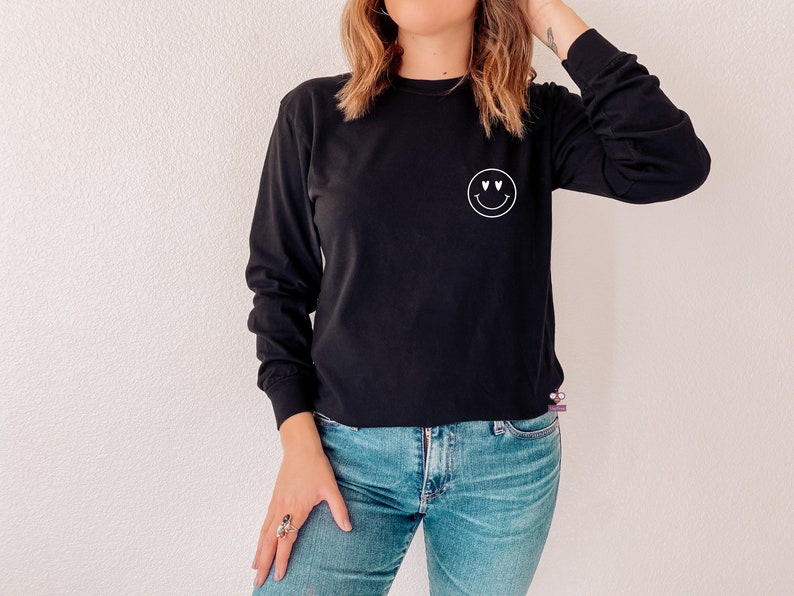 May include: A black long-sleeved shirt with a white smiley face graphic with heart eyes. The shirt is worn by a person with long brown hair and blue jeans.
