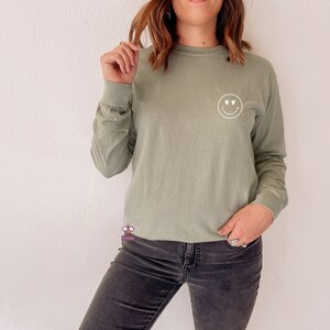 May include: A sage green long-sleeved t-shirt with a white smiley face graphic with hearts for eyes.