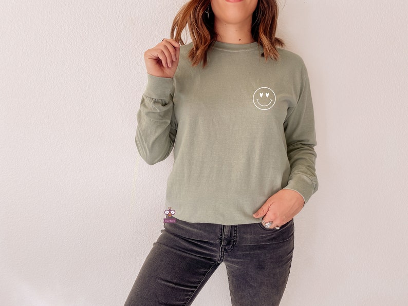 May include: A sage green long-sleeved t-shirt with a white smiley face graphic with hearts for eyes.
