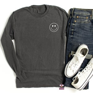 May include: A dark gray long-sleeve t-shirt with a white smiley face with heart eyes graphic on the left chest.
