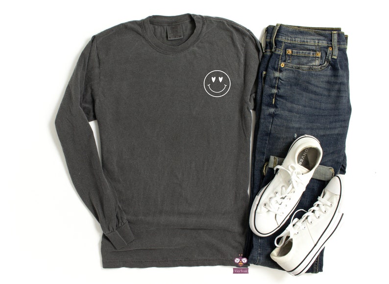 May include: A dark gray long-sleeve t-shirt with a white smiley face with heart eyes graphic on the left chest.