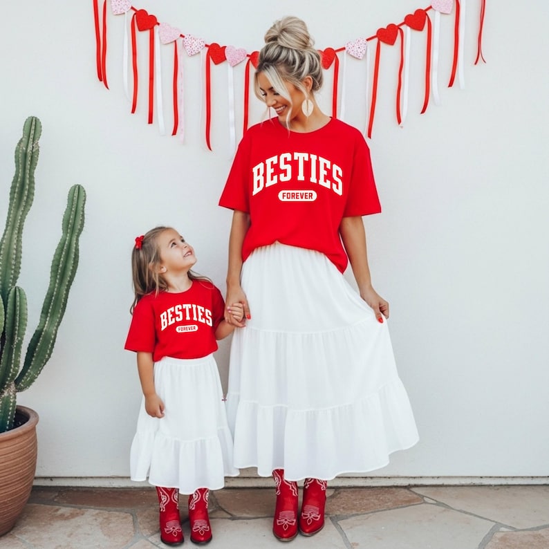 May include: Two people wearing matching red t-shirts with the word "BESTIES" in white, and white skirts. The shirts also have the word "FOREVER" in white. They are holding hands. Red boots complete the outfits. A cactus and Valentine's Day decorations are in the background.