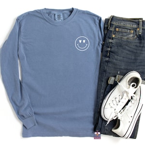 May include: A blue long-sleeve t-shirt with a white smiley face with heart eyes graphic on the front.