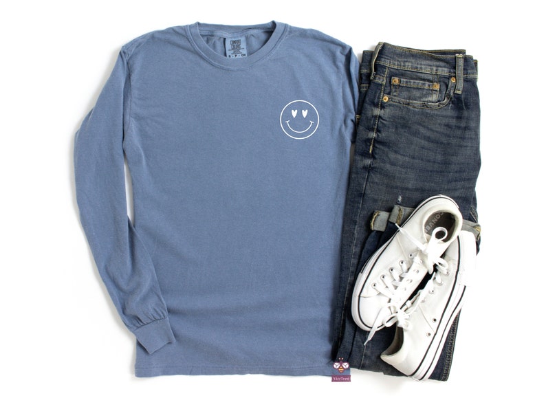 May include: A blue long-sleeve t-shirt with a white smiley face with heart eyes graphic on the front.