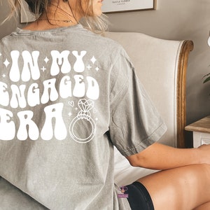 May include: Gray t-shirt with the text "In My Engaged Era" in white retro font with a diamond ring graphic.