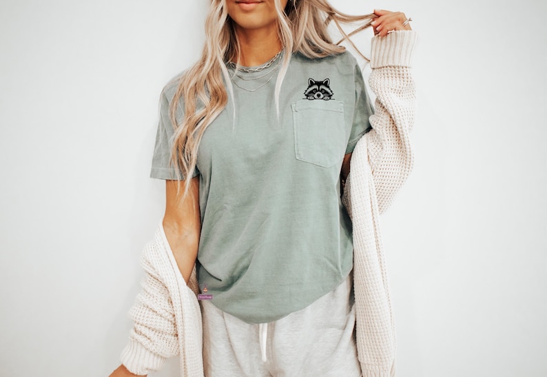 May include: A sage green t-shirt with a black raccoon graphic on the pocket.