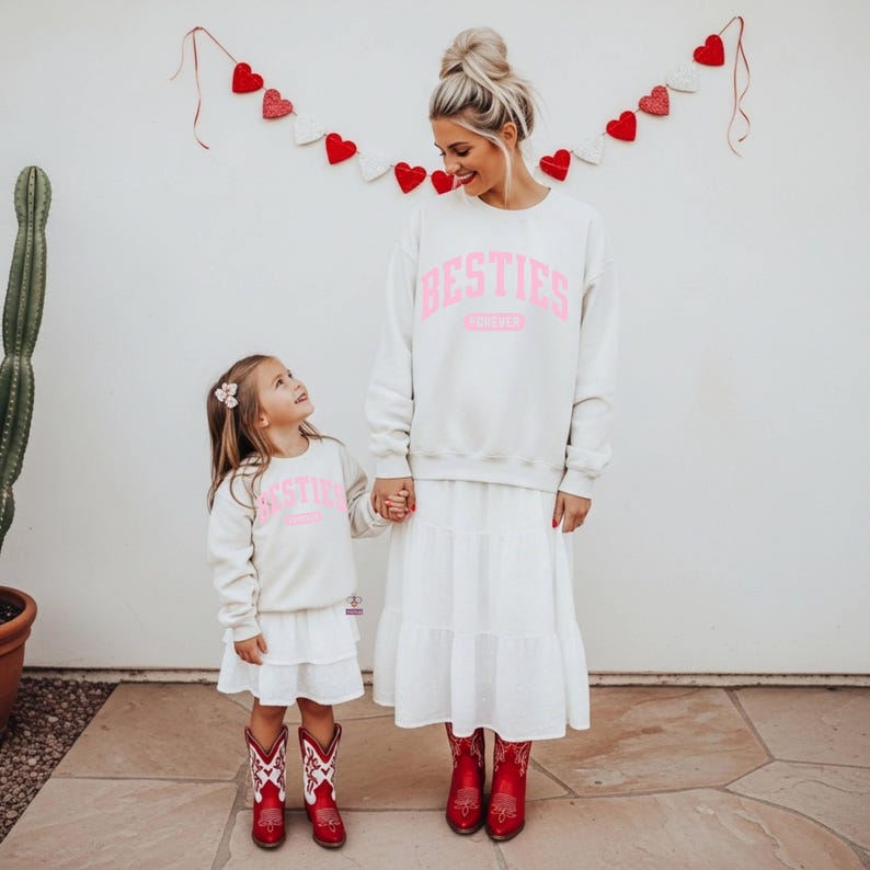 May include: Matching cream-colored sweatshirts with the word "BESTIES" in pink, worn by a woman and a child. The woman wears a white tiered skirt, and both wear red cowboy boots. A heart garland hangs above.