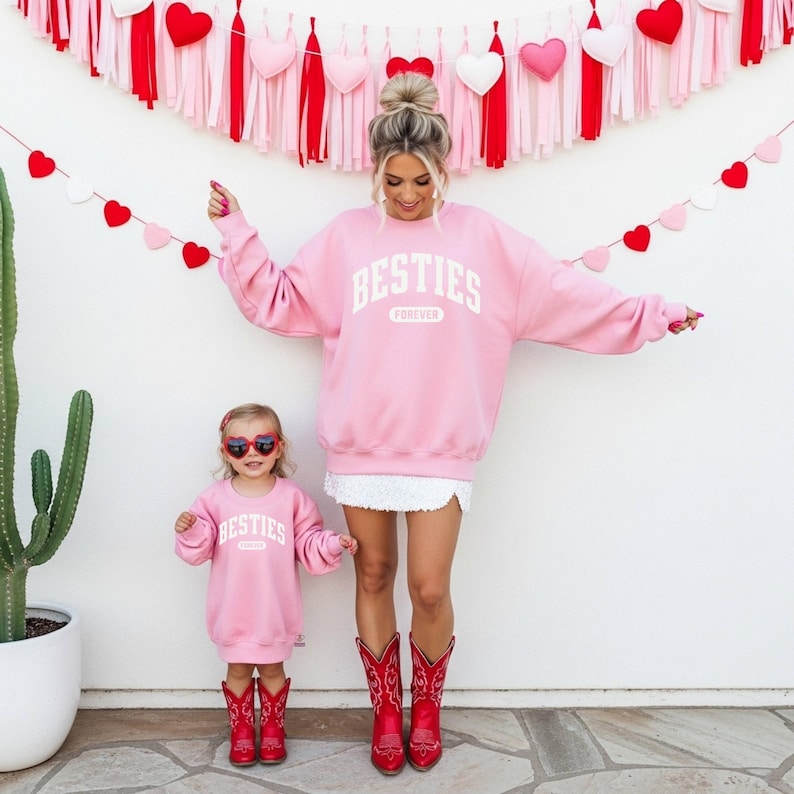 May include: Matching pink sweatshirts with the word "BESTIES FOREVER" in white. The adult is wearing a white skirt and red cowboy boots. The child is wearing heart-shaped sunglasses and red cowboy boots. A cactus and Valentine's Day decorations are in the background.