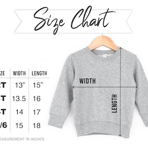May include: A gray sweatshirt displayed on a wooden hanger, with a size chart showing measurements in inches. The chart lists sizes 2T, 3T, 4T, and 5/6, with corresponding width and length measurements. The text "Size Chart" is written above the sweatshirt.