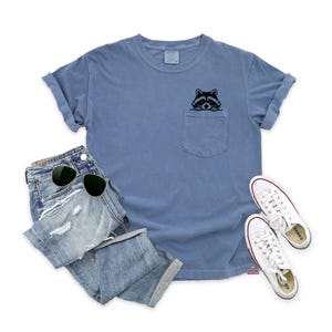 May include: A light blue t-shirt with a pocket featuring a black and white raccoon peeking out. The t-shirt is paired with distressed denim jeans and white sneakers.