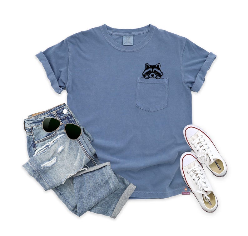 May include: A light blue t-shirt with a pocket featuring a black and white raccoon peeking out. The t-shirt is paired with distressed denim jeans and white sneakers.