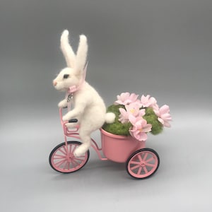 Könnte beinhalten: Ein weißer Filz-Hase fährt auf einem rosa Dreirad, das einen rosa Korb mit rosa Blumen und grünem Moos trägt. Der Hase trägt ein rosa Band. Das Dreirad hat schwarze Räder.