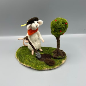 Könnte beinhalten: Eine weiße Filzmaus mit einem braunen Hut und einem orangefarbenen Schal gräbt mit einer kleinen Schaufel ein Loch in den Boden. Im Hintergrund steht ein grüner Filzbaum mit roten Filzbeeren.